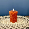 An orange pillar candle with carved leaf patterns, lit with a small flame, sitting on a woven placemat against a blue background.  Pillar Candle available at Quilted Cabin in Airdrie