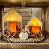 Two house-shaped wooden candle holders with glowing orange pillar candles, arranged on a woven round mat with autumn leaves and a small white pumpkin ornament on a wooden shelf.  Decor items available at Quilted Cabin in Airdrie