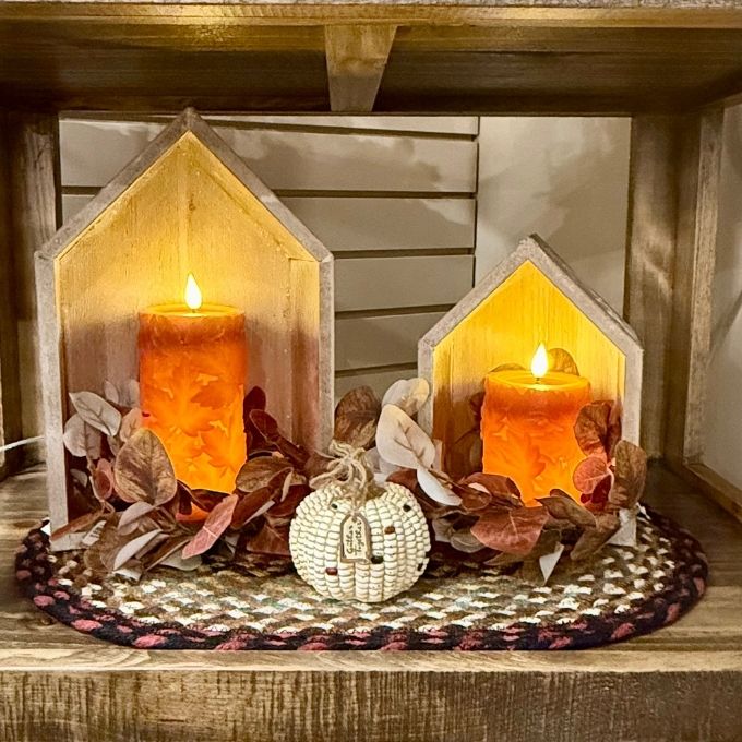 Two house-shaped wooden candle holders with glowing orange pillar candles, arranged on a woven round mat with autumn leaves and a small white pumpkin ornament on a wooden shelf.  Decor items available at Quilted Cabin in Airdrie