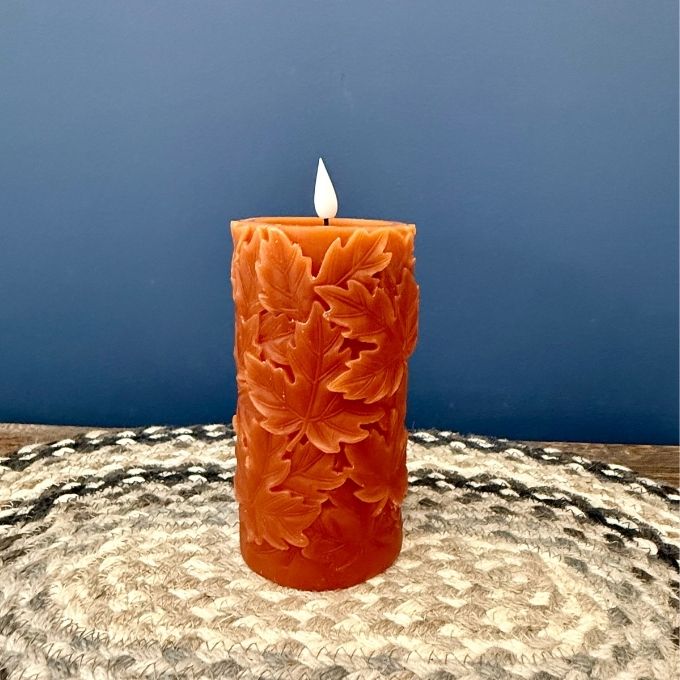 An orange pillar candle with a carved leaf pattern, lit with a white flame, standing on a woven placemat against a blue background.  Two size of candle available at Quilted Cabin in Airdrie