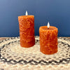 Two orange pillar candles with carved leaf patterns, one tall and one short, displayed on a woven placemat against a blue background. Candles available at Quilted Cabin in Airdrie