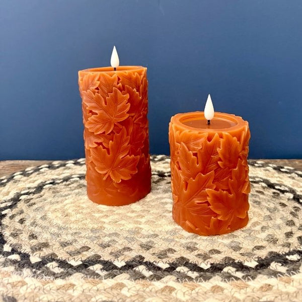 Two orange pillar candles with carved leaf patterns, one tall and one short, displayed on a woven placemat against a blue background. Candles available at Quilted Cabin in Airdrie