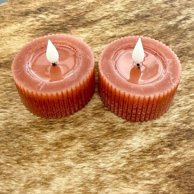 Two small cylindrical orange wax candles with white flame tips, placed side by side on a textured brown surface.  Tealights available at Quilted Cabin in Airdrie