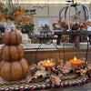 A styled autumn tabletop arrangement with a stacked three-bump pumpkin on the left, two black metal lanterns holding lit orange candles, and scattered faux leaves on a woven runner. The scene is set in a rustic indoor shop or home decor space with additional decor visible in the background.  All Decor items available at Quilted Cabin in Airdrie