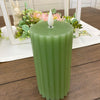 Green candle with a white wick on a wooden surface with flowers in the background.  Sage Green Ridged LED Pillar Timer Candle available at Quilted Cabin Home Decor.