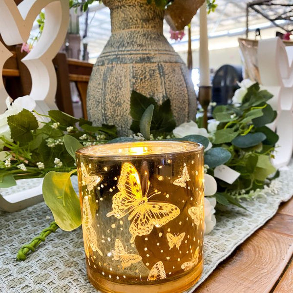 Decorative candle with butterfly design on a table with flowers and a vase. Golden Butterfly Glass Jar LED Timer Candle available at Quilted Cabin Home Decor.