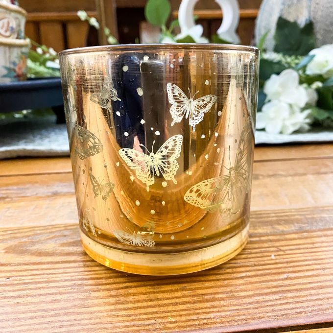 Glass candle holder with butterfly designs on a wooden surface.  Golden Butterfly Glass Jar LED Timer Candle available at Quilted Cabin Home Decor.