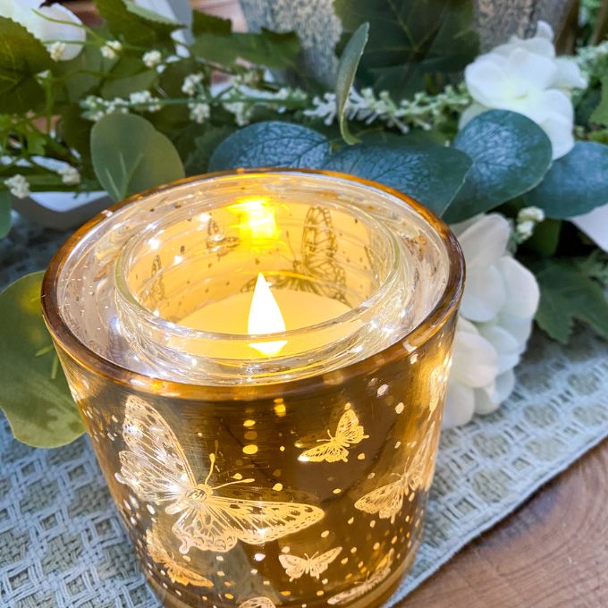Candle in a glass holder with butterfly design, surrounded by flowers and greenery.  Golden Butterfly Glass Jar LED Timer Candle available at Quilted Cabin Home Decor.