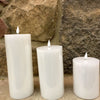 Three white candles of different sizes against a stone wall. White LED Timer Pillar Candles - Three Sizes available at Quilted Cabin Home Decor.