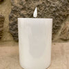 White candle with a flame against a stone wall background. White LED Timer Pillar Candles - Three Sizes available at Quilted Cabin Home Decor.