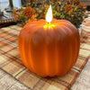 Orange pumpkin-shaped candle with a lit wick on a checkered tablecloth. Orange LED Pumpkin Candle available at Quilted Cabin Home Decor.