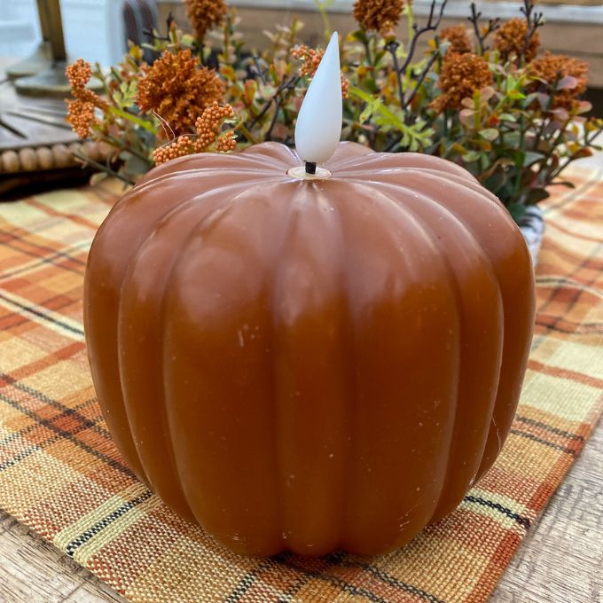 Pumpkin-shaped candle with a white wick on a plaid surface with autumn decorations. Orange LED Pumpkin Candle available at Quilted Cabin Home Decor.