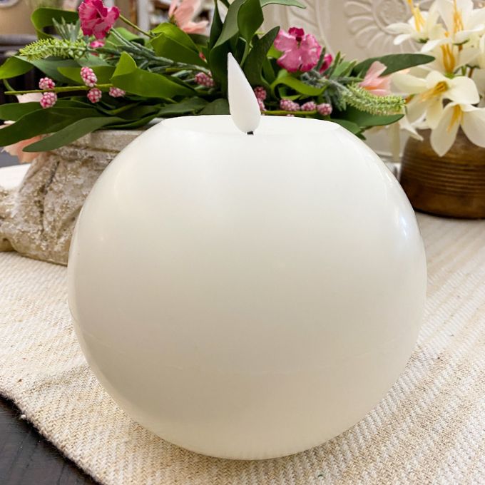 White spherical candle on a textured surface with flowers in the background. White Round LED Timer Candle available at Quilted Cabin Home Decor.