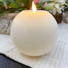White spherical candle with a flame on a textured surface with flowers in the background. White Round LED Timer Candle available at Quilted Cabin Home Decor.