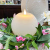 Candle surrounded by greenery and pink flowers on a decorative plate. White Round LED Timer Candle available at Quilted Cabin Home Decor.