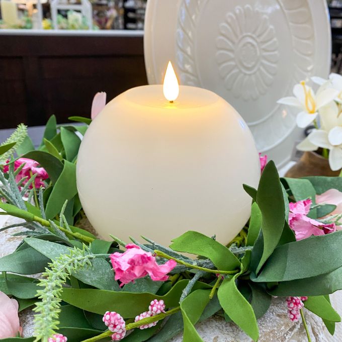 Candle surrounded by greenery and pink flowers on a decorative plate. White Round LED Timer Candle available at Quilted Cabin Home Decor.