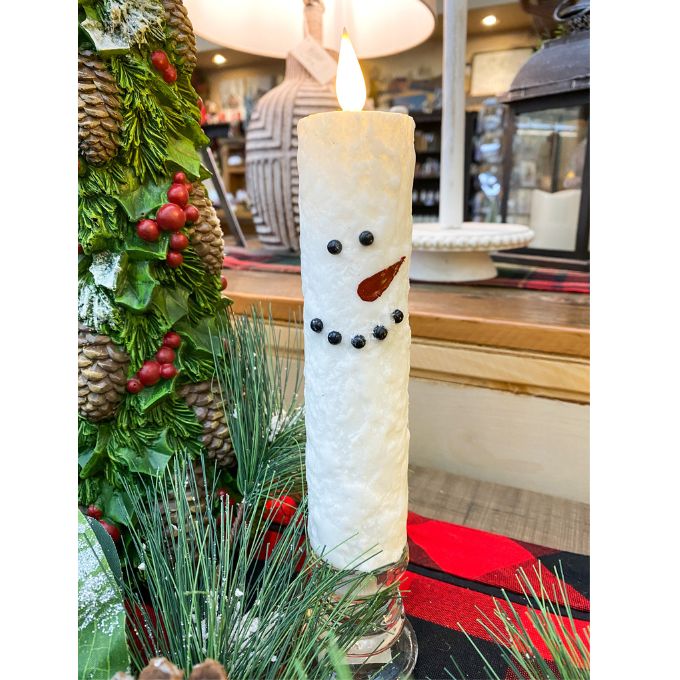 Decorative candle shaped like a snowman with greenery and a blurred indoor background. Snowman LED Taper Timer Candle available at Quilted Cabin Home Decor.