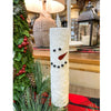 Decorative candle shaped like a snowman with greenery and a lamp in the background. Snowman LED Taper Timer Candle available at Quilted Cabin Home Decor.