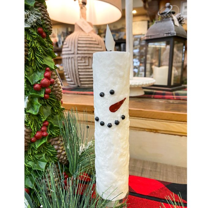 Decorative candle shaped like a snowman with greenery and a lamp in the background. Snowman LED Taper Timer Candle available at Quilted Cabin Home Decor.