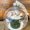 Clear glass ornament with a miniature Christmas scene inside, featuring a tree and figures, on a wooden surface. Ball Ornaments with LED Candles - Three Styles available at Quilted Cabin Home Decor.