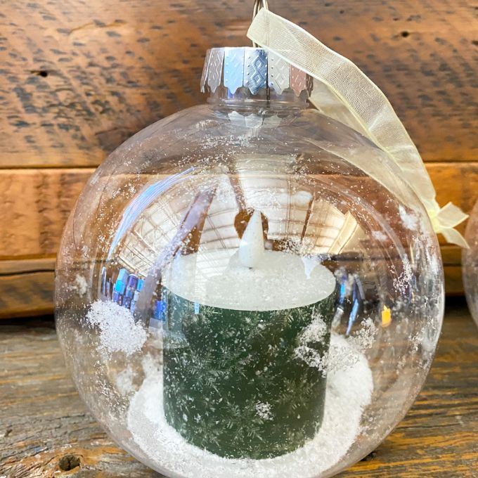 Clear glass ornament with a miniature Christmas scene inside, featuring a tree and figures, on a wooden surface. Ball Ornaments with LED Candles - Three Styles available at Quilted Cabin Home Decor.
