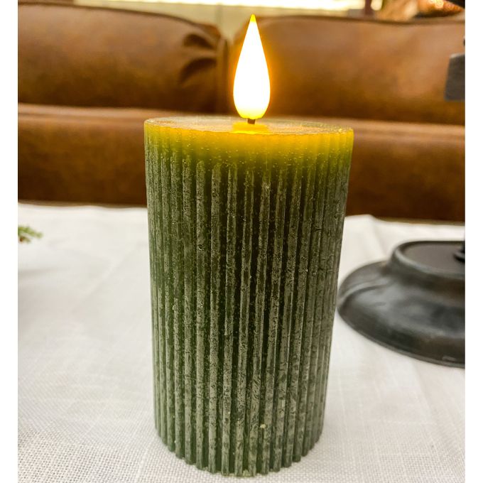 Green textured candle with a flame on a white surface with a brown leather couch in the background. Green Ridged LED 4" Votive Candle available at Quilted Cabin Home Decor.