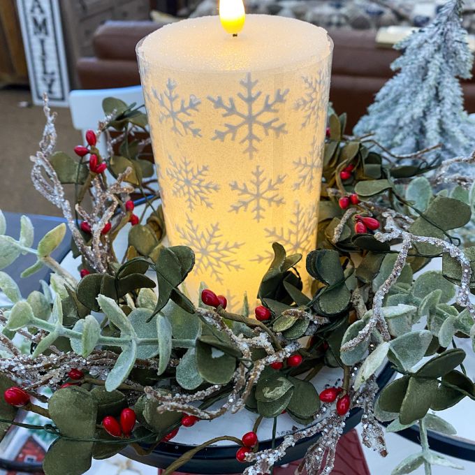 Decorative candle with snowflake pattern surrounded by greenery and berries on a table.  White Snowflake LED 5" Pillar Candle available at Quilted Cabin Home Decor.
