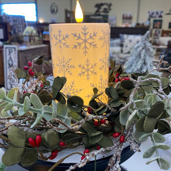 Decorative candle with snowflake pattern surrounded by greenery and red berries in a festive setting. White Snowflake LED 5" Pillar Candle available at Quilted Cabin Home Decor.