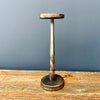 A rustic metal T-shaped candle holder with a twisted shaft and round base, displayed on a wooden surface against a dark blue-gray wall.  Two size of candle holder available at Quilted Cabin in Airdrie