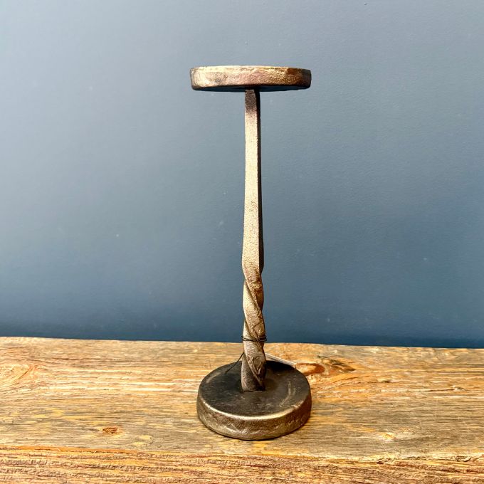 A rustic metal T-shaped candle holder with a twisted shaft and round base, displayed on a wooden surface against a dark blue-gray wall.  Two size of candle holder available at Quilted Cabin in Airdrie