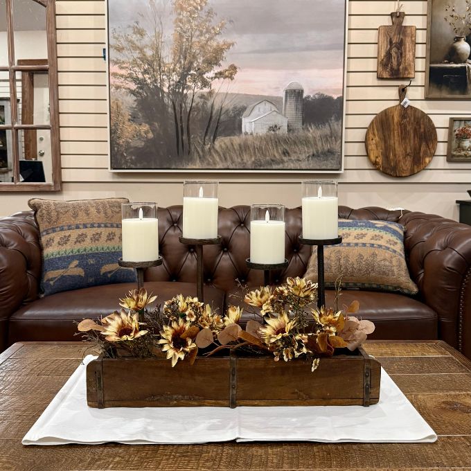 A styled living room scene featuring a brown leather sofa with patterned throw pillows, a large framed landscape painting on the wall, and a rustic wooden tray filled with dried sunflowers and autumn leaves on a white runner on the coffee table.  All items available at Quilted Cabin in Airdrie