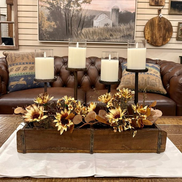 A rustic fall-themed centerpiece in a wooden tray, featuring dried sunflowers, eucalyptus leaves, and a black metal three-tier candle holder with four white pillar candles, displayed on a white cloth in front of a brown leather sofa.  Both sizes of candle holders available at Quilted Cabin in Airdrie
