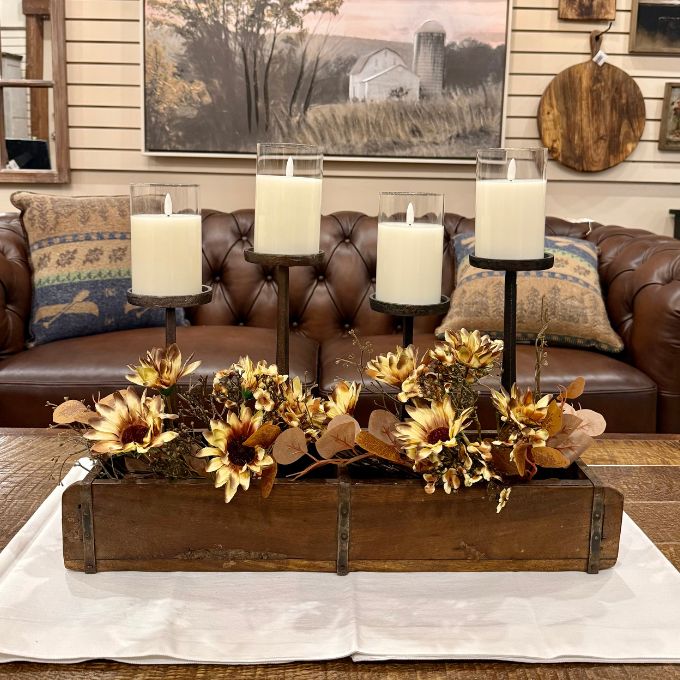 A rustic fall-themed centerpiece in a wooden tray, featuring dried sunflowers, eucalyptus leaves, and a black metal three-tier candle holder with four white pillar candles, displayed on a white cloth in front of a brown leather sofa.  Both sizes of candle holders available at Quilted Cabin in Airdrie