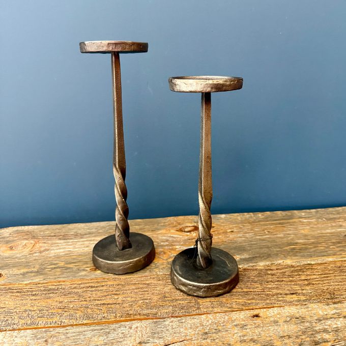 Two rustic metal candlestick holders with twisted shafts and round bases, displayed on a wooden surface against a blue wall.  Candle holders available at Quilted Cabin in Airdrie