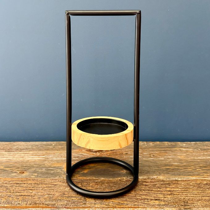 A modern floating candle holder featuring a black metal frame and a light natural wood ring floating inside a clear glass container, set on a rustic wooden table against a slate blue background.  Candle holder available at Quilted Cabin in Airdrie