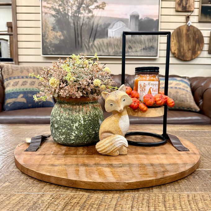 A rustic wooden oval serving board on a table, styled with a green speckled vase of dried flowers, a small fox figurine, and a black metal two-tier stand holding a jar of honey and orange berries, set in a cozy living room environment.  All items available at Quilted Cabin in Airdrie