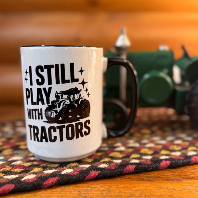 Mug with 'I Still Play with Tractors' text and graphic on a wooden surface with a blurred background. Available at Quilted Cabin Home Decor.