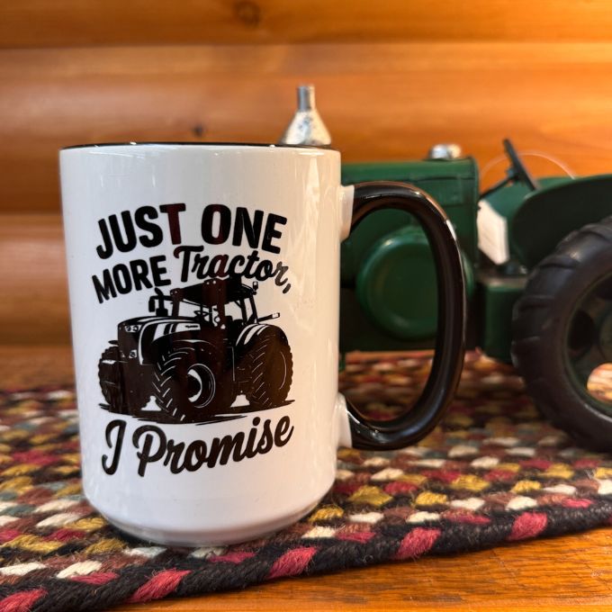 White mug with tractor design and text on a wooden surface with a toy tractor in the background. Just One more Tractor I promise Mug is available at Quilted Cabin Home Decor.
