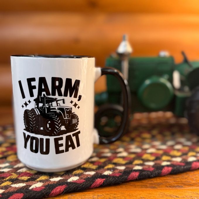White mug with 'I Farm, You Eat' text and tractor graphic on a wooden surface with a blurred background. available at Quilted CAbin Home Decor.