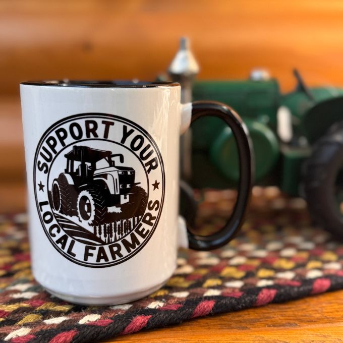 White mug with black handle featuring 'Support Your Local Farmers' design on a wooden surface with a blurred background. Available at Quilted Cabin Home Decor.