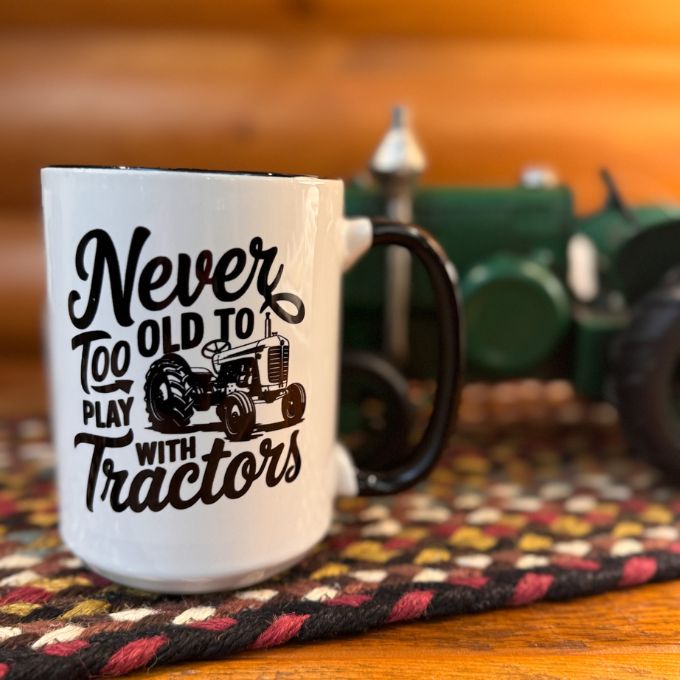 White mug with 'Never Too Old to Play with Tractors' text and graphic on a wooden surface with a blurred tractor in the background. Available at Quilted Cabin Home Decor.