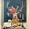 A framed wall art print depicting a deer with large antlers decorated with flowers and birds, displayed above a small white vintage truck and two black candlesticks on a tabletop. All items available at Quilted Cabin in Airdrie