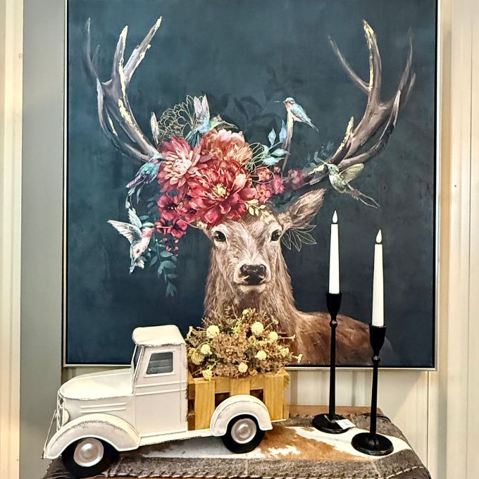 A framed wall art print depicting a deer with large antlers decorated with flowers and birds, displayed above a small white vintage truck and two black candlesticks on a tabletop. All items available at Quilted Cabin in Airdrie