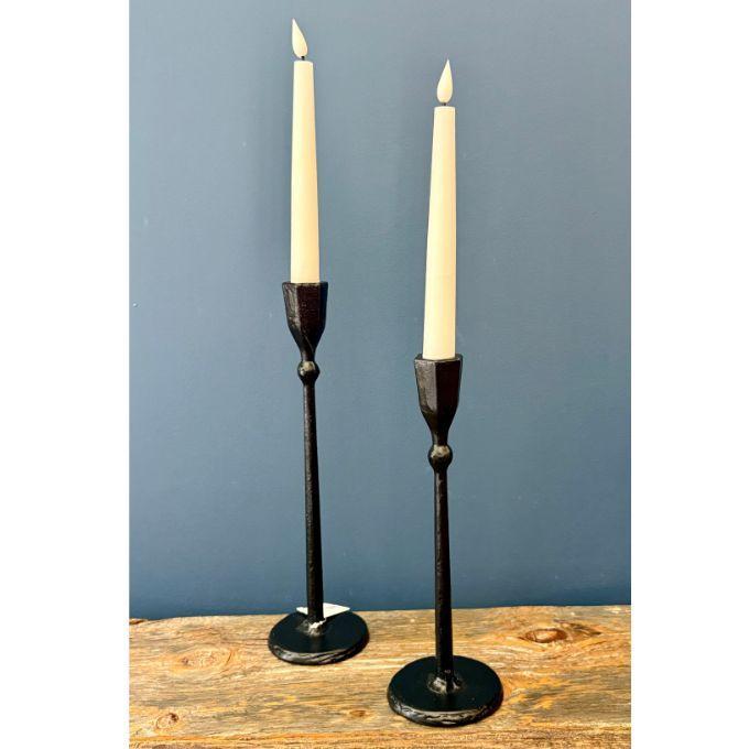 Two black metal taper candlesticks holding cream-colored candles, displayed on a rustic wooden surface against a solid blue-gray background. Candle sticks come in 2 Sizes available at Quilted Cabin in Airdrie