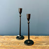 Two black cast iron candlesticks with a rustic, weathered finish, standing on a wooden surface against a slate blue background. Available at Quilted Cabin in Airdrie