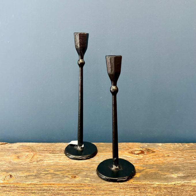 Two black cast iron candlesticks with a rustic, weathered finish, standing on a wooden surface against a slate blue background. Available at Quilted Cabin in Airdrie