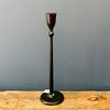 A dark cast iron candlestick holder with a textured finish, standing on a rustic wooden surface against a solid gray background. Two sizes available at Quilted Cabin in Airdrie