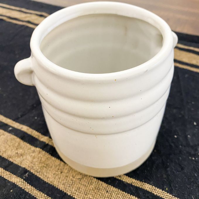 White ceramic cup with handles on a striped fabric background. White Farmhouse-Styled Ceramic Jar available at Quilted Cabin Home Decor.