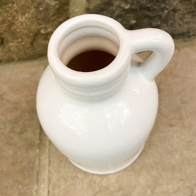 White ceramic pitcher against a stone wall background. White Porcelain Jug-Style Vase available at Quilted Cabin Home Decor. 