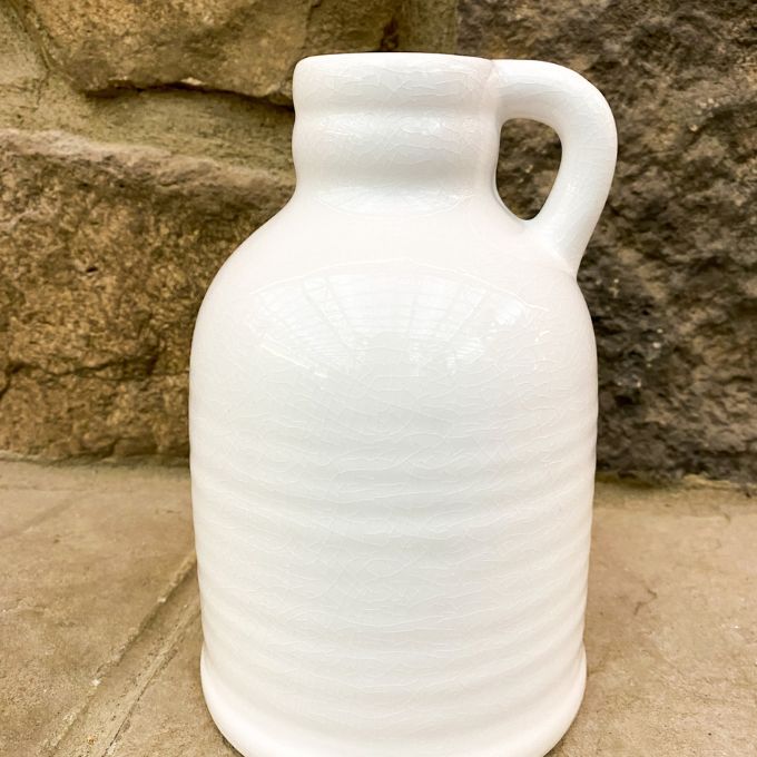 White ceramic jug with a handle against a stone wall. White Porcelain Jug-Style Vase available at Quilted Cabin Home Decor. 